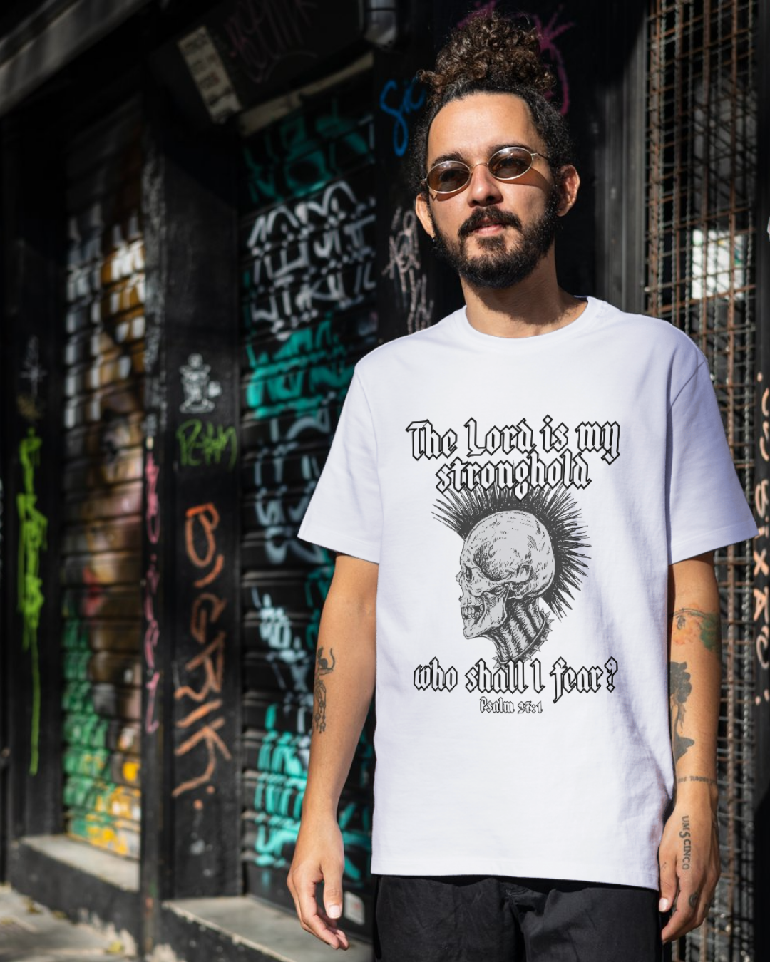 Man wearing a white t-shirt with a graphic design and text, standing in front of a graffiti-covered wall.